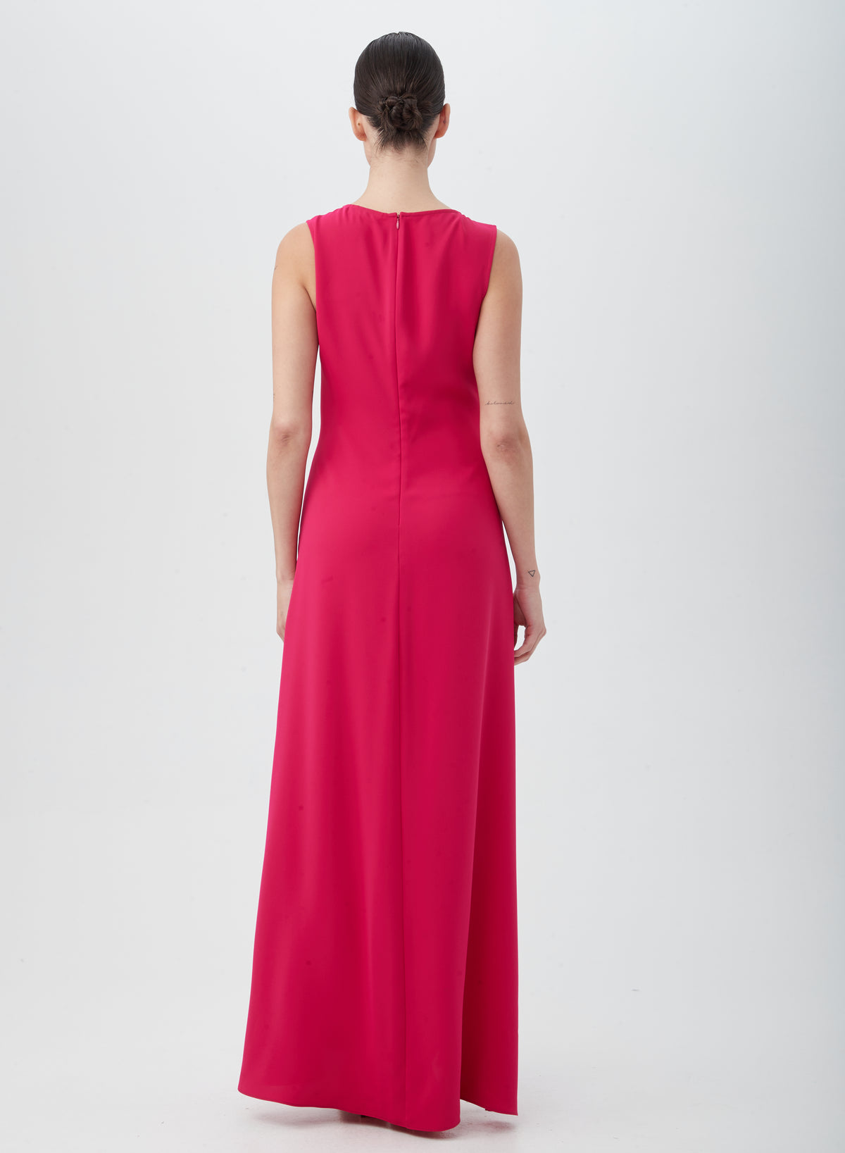 A woman with dark hair in a bun faces away, wearing the MYTHICAL DRESS—a sleeveless, floor-length bright pink dress made from 100% polyester. She stands against a plain white background.