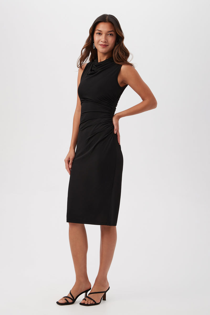 A woman poses against a white background wearing the Spruce Sleeveless Slinky Jersey Midi Dress With Cowl Neck. The black, knee-length dress features ruched details; she styles it with black heeled sandals and wears her long hair down, smiling softly.