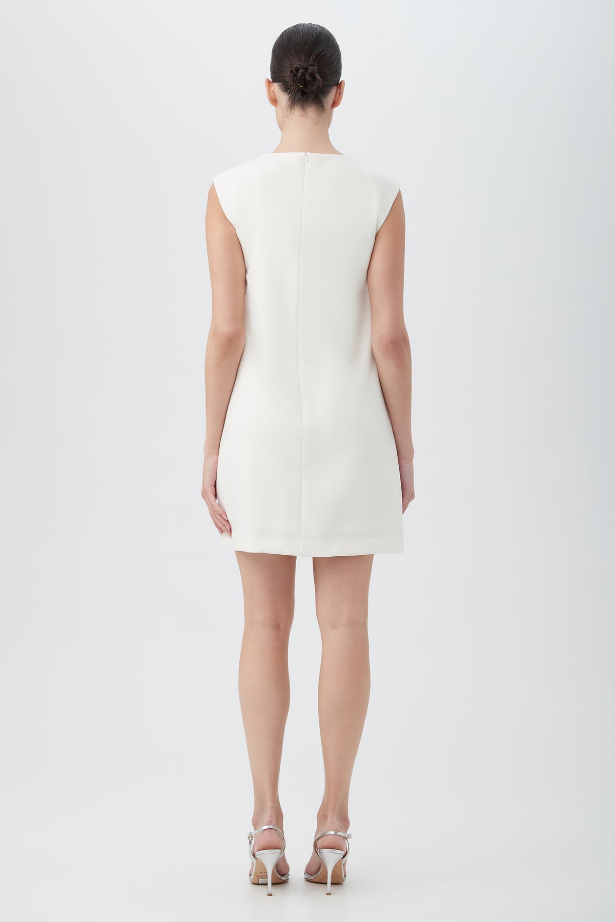 A woman faces away, wearing the Ruth Sleeveless Shift Dress With Decorative Front Detail in white and silver high-heeled sandals. Her hair is neatly pulled back as she poses against a plain light background.