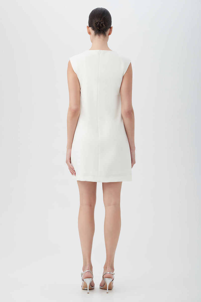 A woman faces away, wearing the Ruth Sleeveless Shift Dress With Decorative Front Detail in white and silver high-heeled sandals. Her hair is neatly pulled back as she poses against a plain light background.