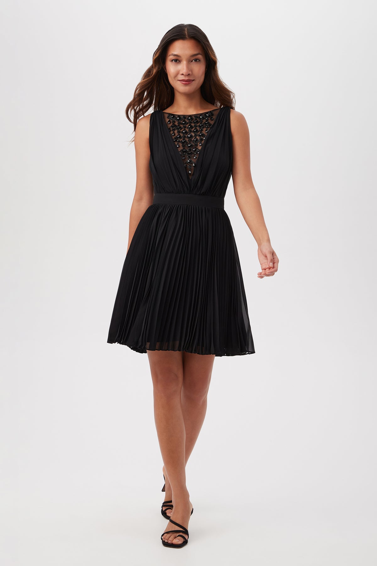 A woman with long brown hair smiles slightly as she walks forward in the Zathar Sleeveless Dress With Jeweled Front And Accordion Pleats, black strappy heels, against a plain white background. The dress is black and knee-length.
