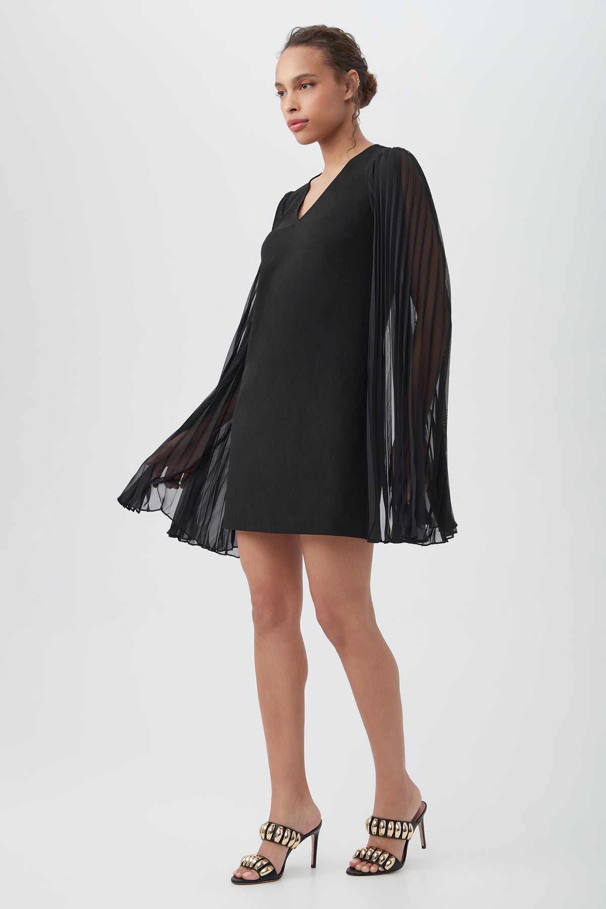 A woman poses in the Celestina Crepe Knit Sheath Dress, featuring a black V-neck mini silhouette with long sheer split sleeves, paired with open-toe heeled sandals adorned with gold chain accents against a plain light background.