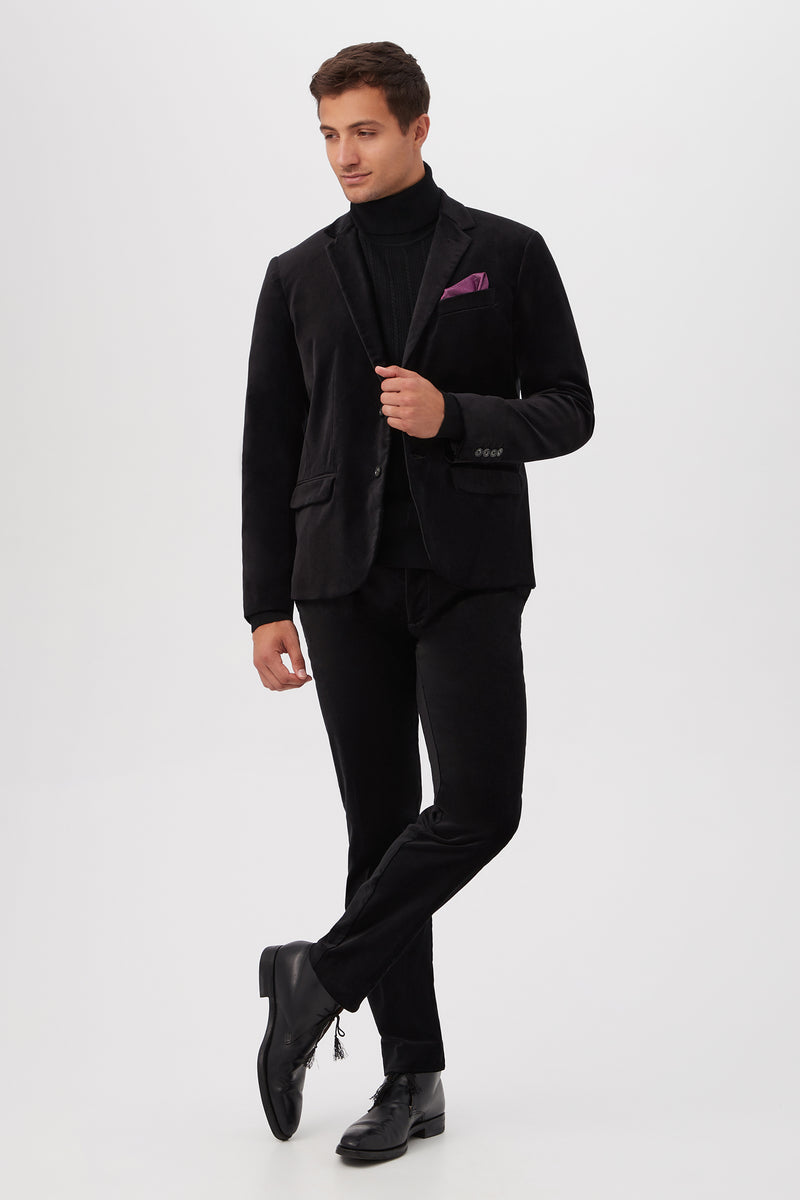 A man stands against a white background wearing the Clyde Slim Trouser in Black Velveteen, paired with a black blazer, black turtleneck, black dress shoes, and a purple pocket square. He looks to the side with one leg crossed in front.