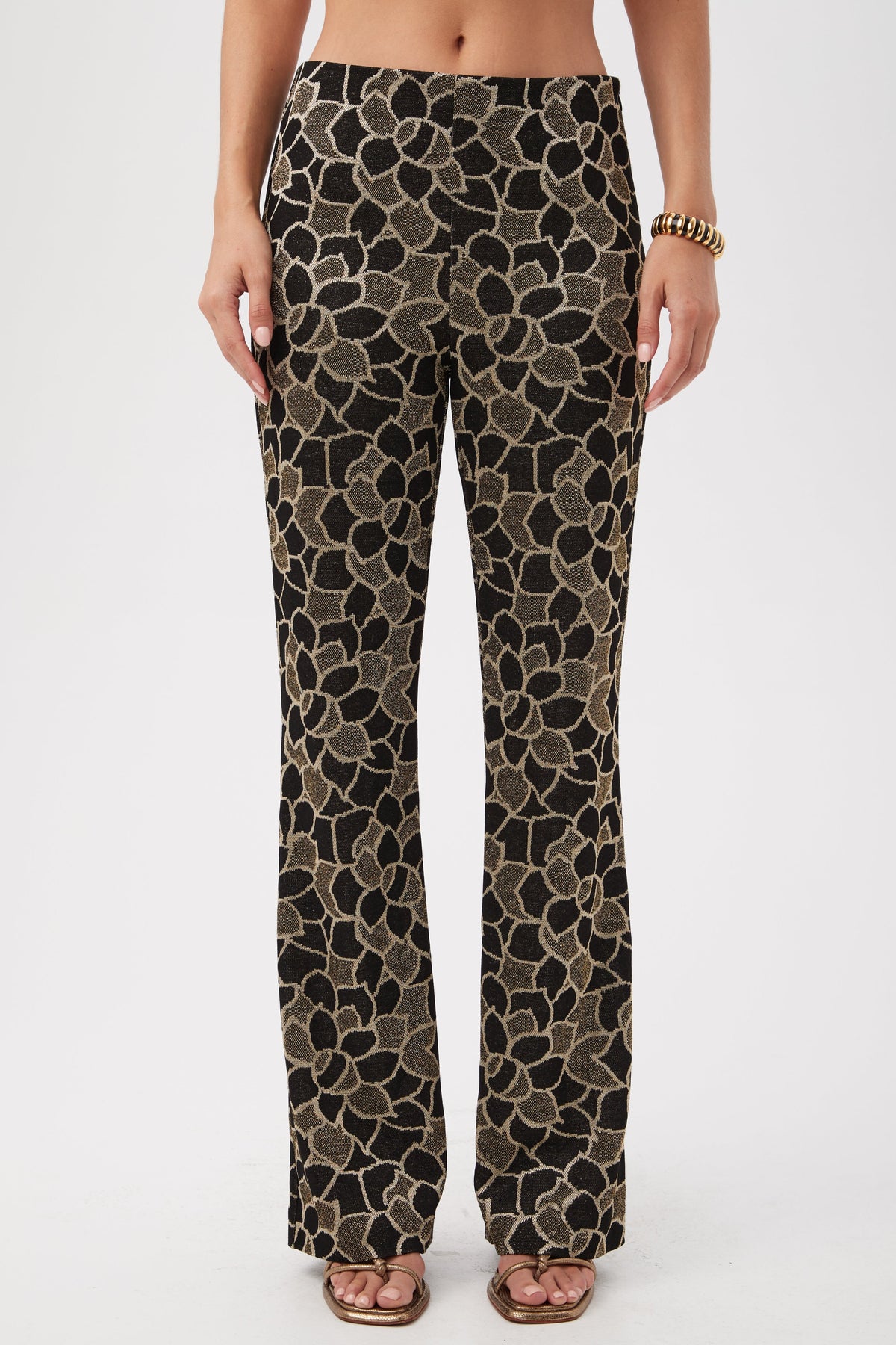 A person wearing Jacoba Metallic Knit Jacquard Bootcut Pants in multi, featuring a bold floral pattern, stands in sandals and a gold bracelet against a plain white background.
