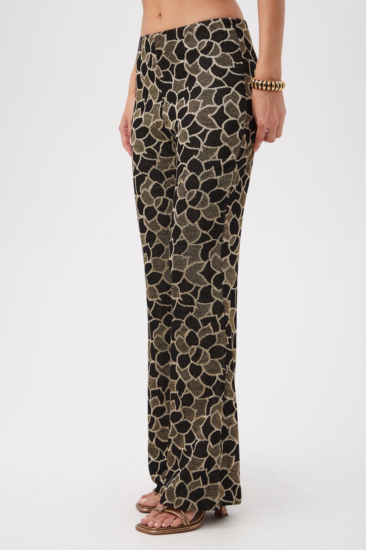 Someone stands against a plain white background wearing Jacoba Metallic Knit Jacquard Bootcut Pants, featuring a bold black and beige floral pattern, styled with open-toe sandals and a chunky gold bracelet.
