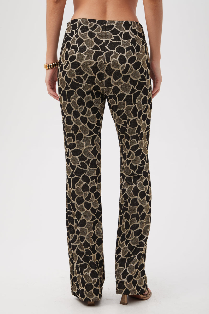 A person stands facing away, wearing Jacoba Metallic Knit Jacquard Bootcut Pants featuring a bold black and gold floral pattern, plus a gold bracelet, against a plain white background.