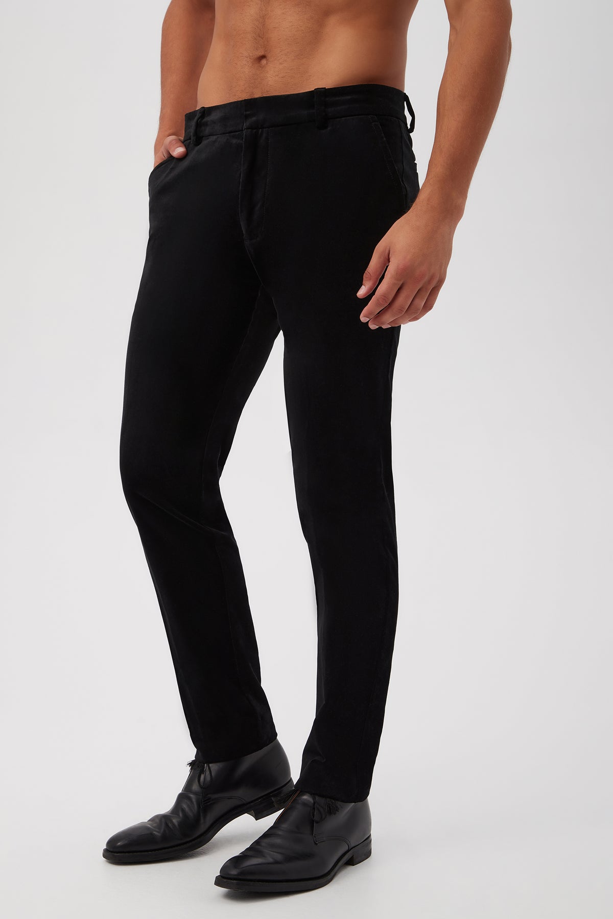 A man models the Clyde Slim Trouser in Black Velveteen, paired with black dress shoes, standing hands-in-pockets against a plain white background with a bare upper body.