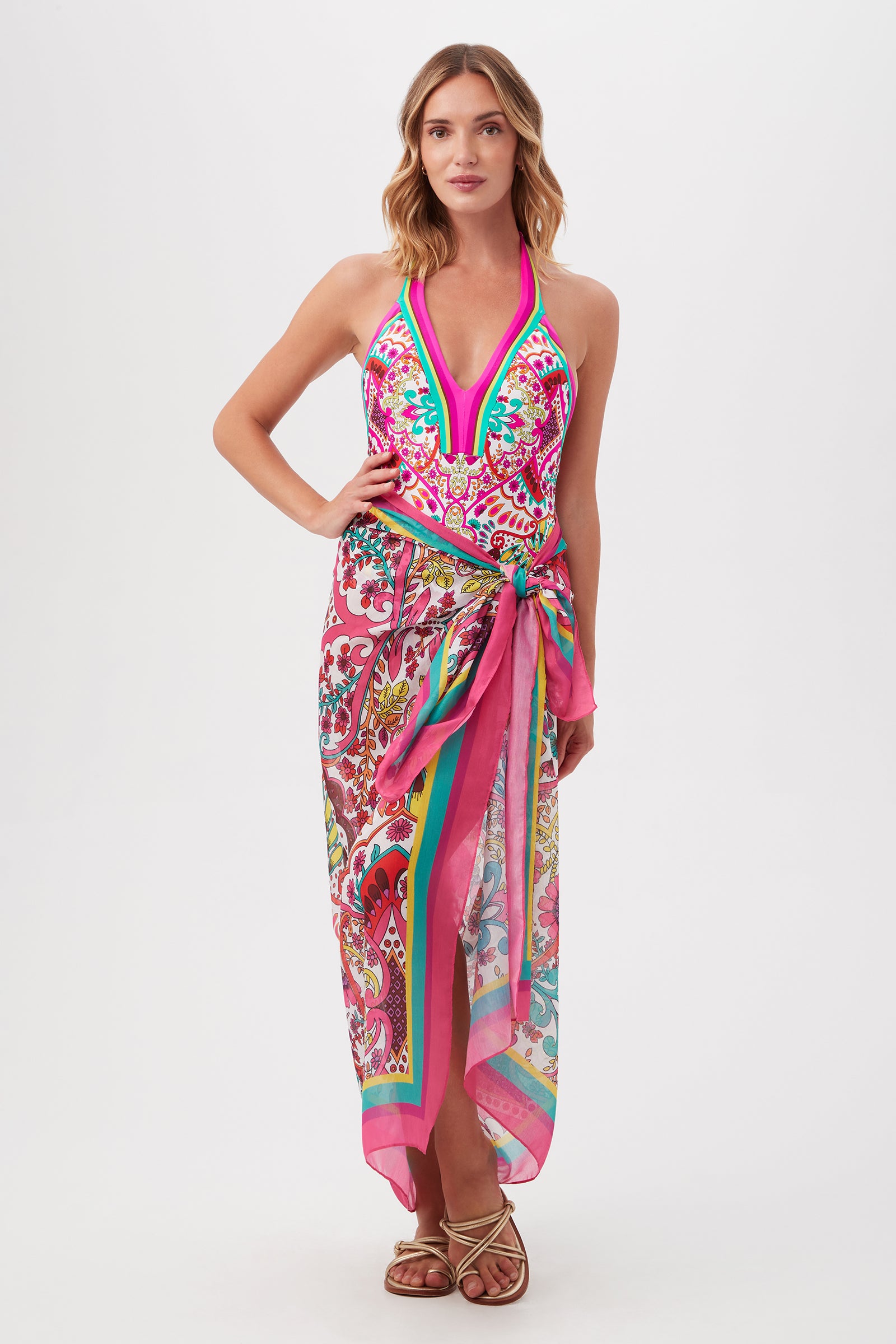 A woman stands barefoot against a white background, her loose waves perfectly styled, wearing the Hawaiian Garden Pareo Swim Cover-Up—a colorful, patterned halter dress with a pink waistband and side slit for a resort-ready look.