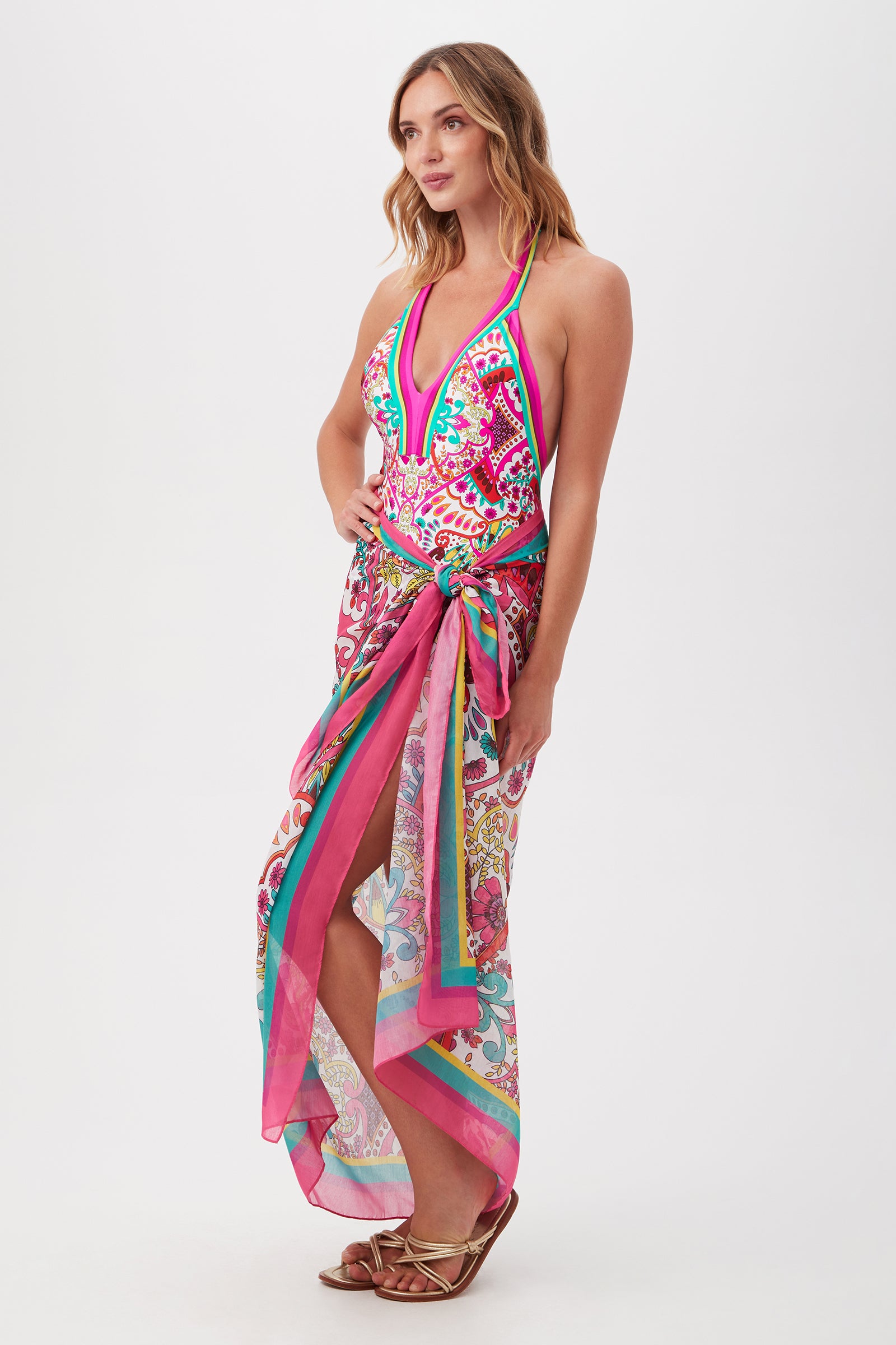 A woman models the Hawaiian Garden Pareo Swim Cover-Up, featuring a vibrant pink and green halter-neck design with a deep neckline and waist sash—ideal as a resort-ready sarong. She pairs it with sandals against a plain white background.