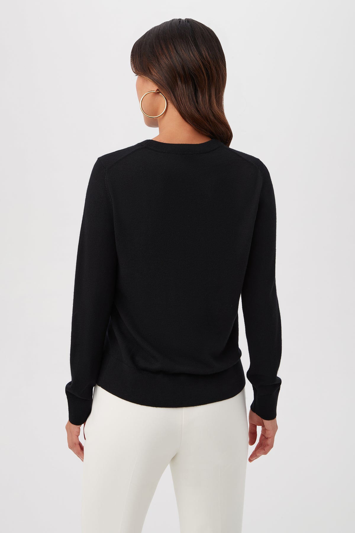 A woman with medium-length brown hair and large hoop earrings, wearing the Gold Heart Merino Wool Crewneck Sweater and white pants, stands facing away from the camera against a plain white background.
