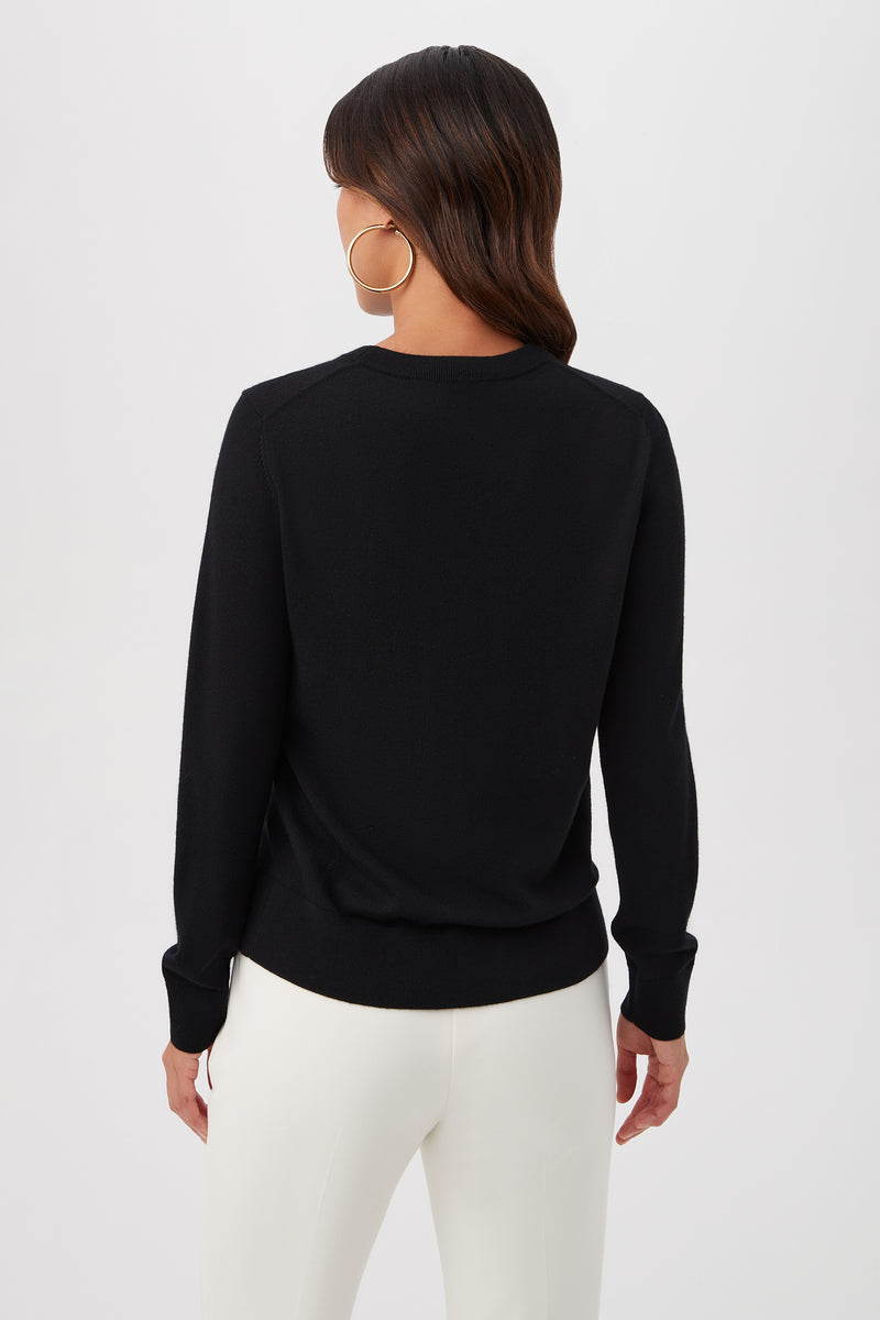 A woman with medium-length brown hair and large hoop earrings, wearing the Gold Heart Merino Wool Crewneck Sweater and white pants, stands facing away from the camera against a plain white background.