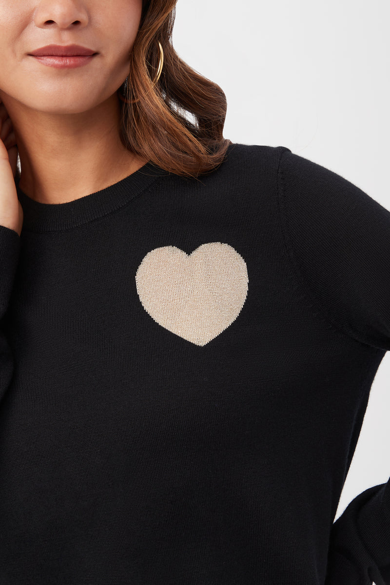 A woman, visible from the shoulders up, smiles softly with one hand near her neck while wearing the Gold Heart Merino Wool Crewneck Sweater featuring a beige heart design on the chest.
