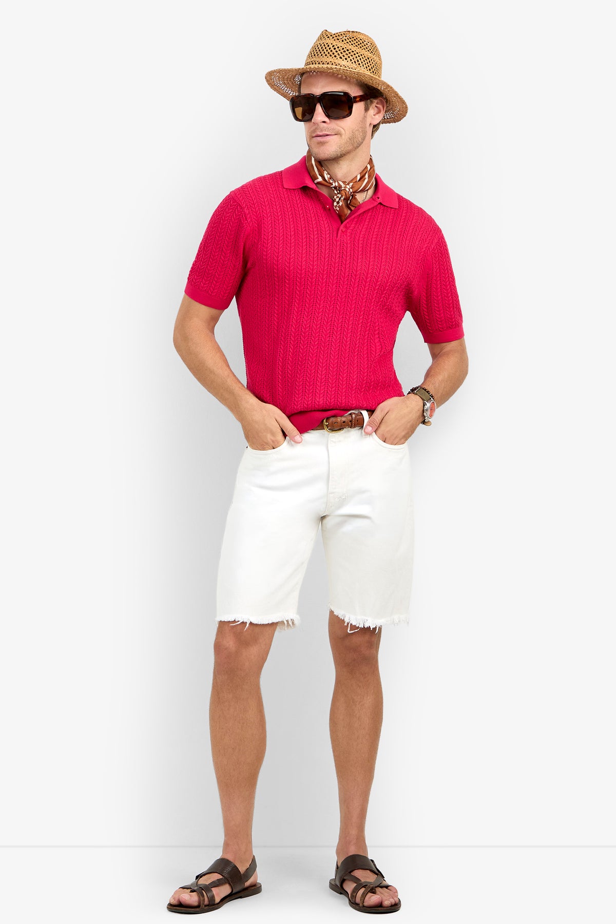 Man wears the Maxton Short-Sleeve Polo - Cotton Modal in red, white frayed shorts, brown sandals, straw hat, sunglasses, and a neck scarf with hands in pockets against a plain white background.