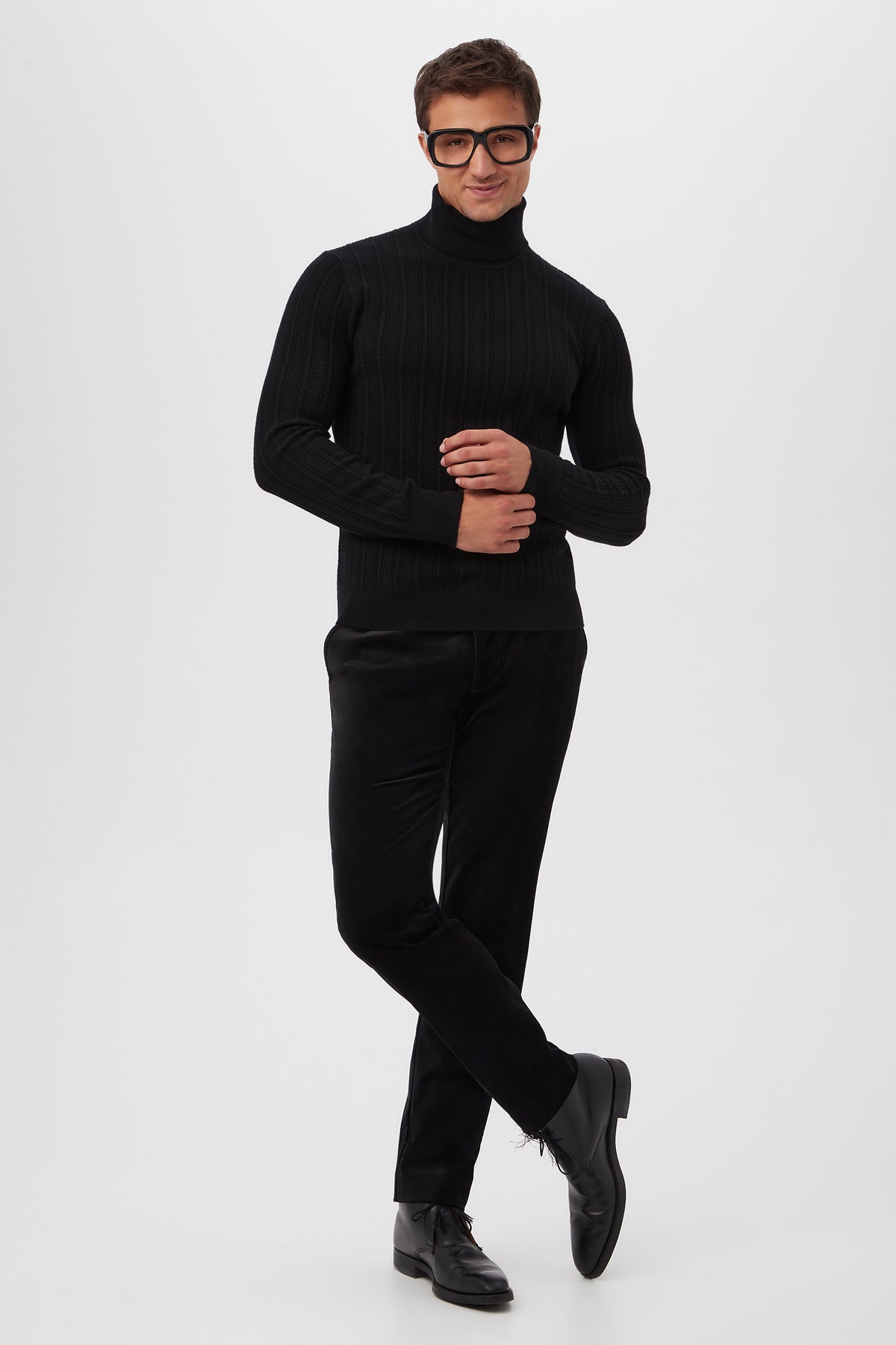 A man poses confidently in the Oberon Turtleneck Sweater - Merino Wool, styled with black pants and shoes. He wears large, dark-rimmed glasses and smiles slightly against a plain white background, standing with one leg crossed and hands together.