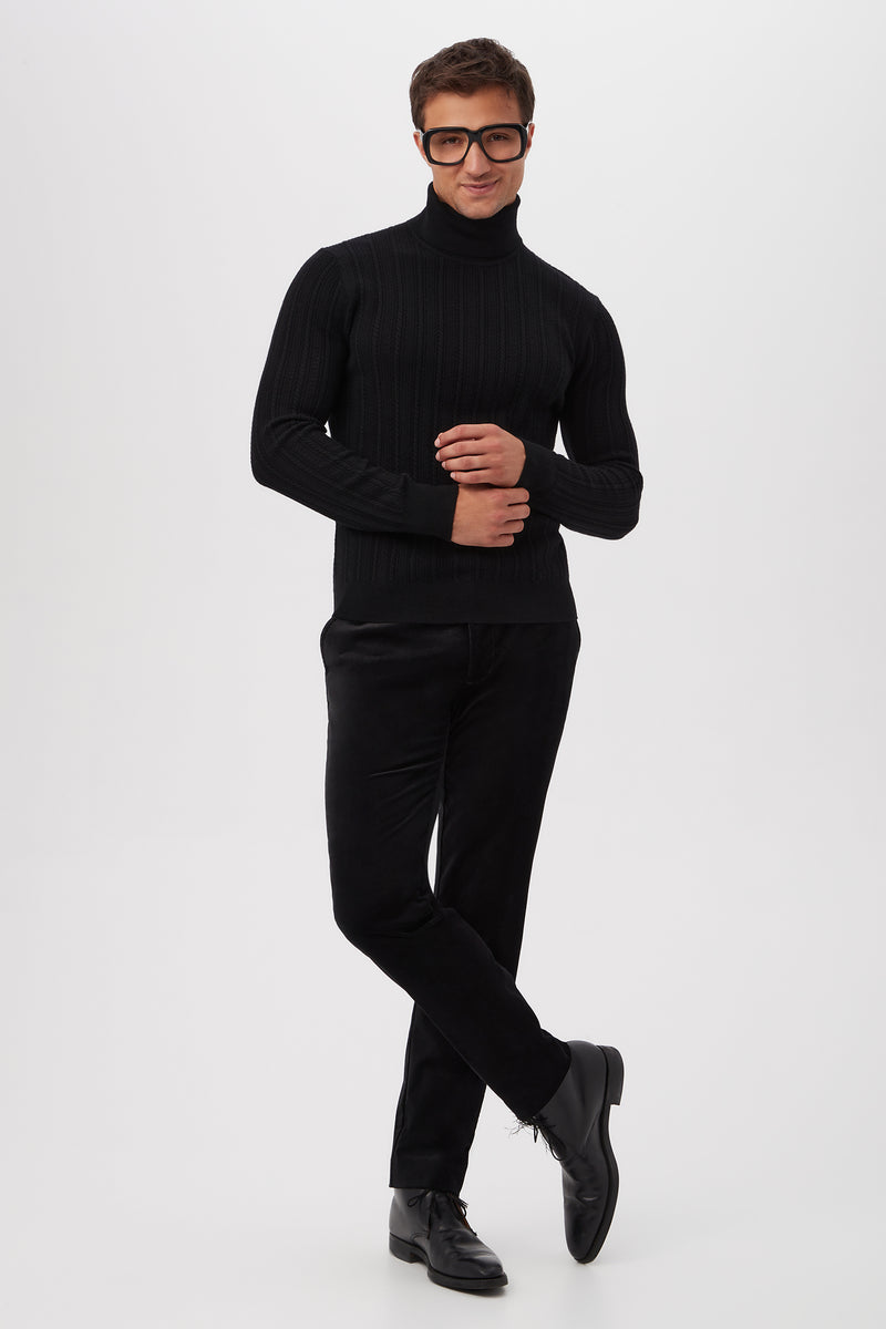 A man poses confidently in the Oberon Turtleneck Sweater - Merino Wool, styled with black pants and shoes. He wears large, dark-rimmed glasses and smiles slightly against a plain white background, standing with one leg crossed and hands together.