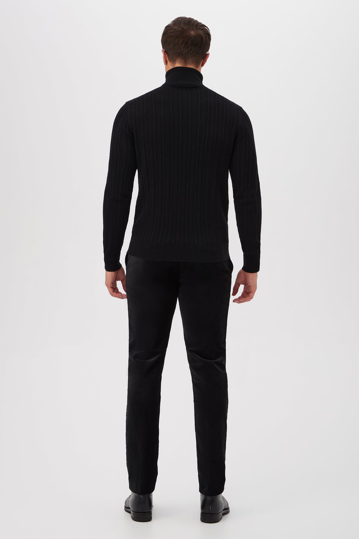 A man faces away from the camera against a white background, wearing the Oberon Turtleneck Sweater - Merino Wool in black, paired with black pants and shoes.