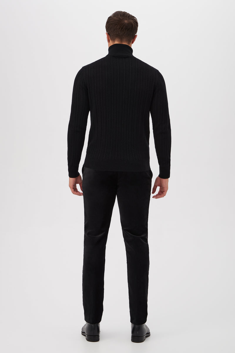 A man faces away from the camera against a white background, wearing the Oberon Turtleneck Sweater - Merino Wool in black, paired with black pants and shoes.