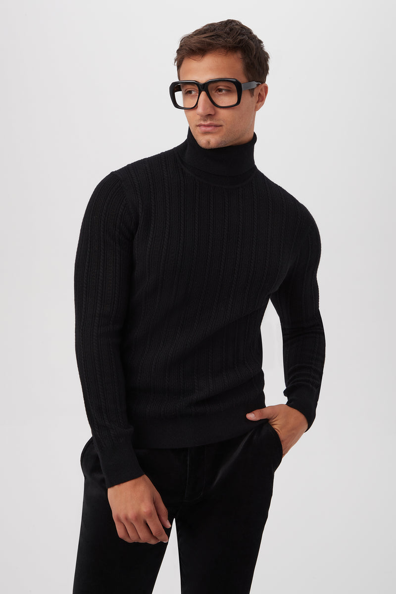 A man stands against a plain white background, wearing the black Oberon Turtleneck Sweater made from merino wool and large black glasses, with one hand in his pocket and looking slightly to the side.