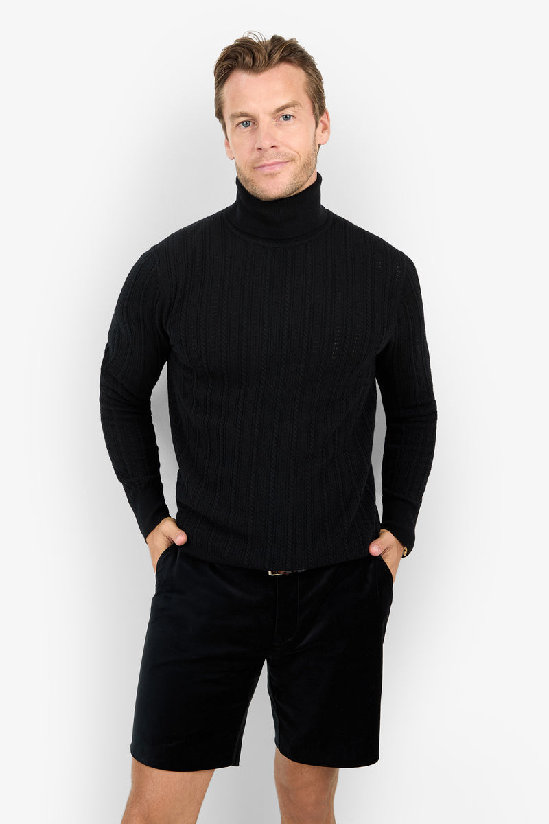 Man wearing the Oberon Turtleneck Sweater - Merino Wool and black shorts, standing with hands in pockets, facing the camera against a plain white background.