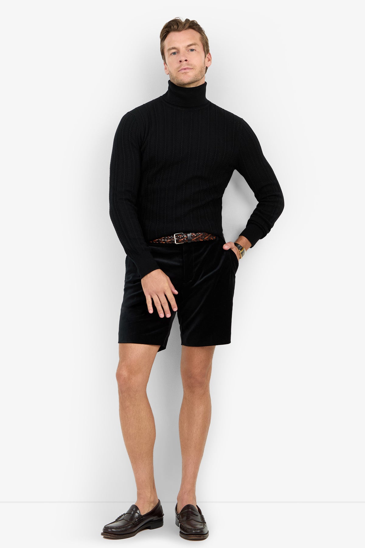 A man poses against a white background wearing the Oberon Turtleneck Sweater - Merino Wool, black shorts, a braided belt, brown loafers, and a wristwatch with one hand resting on his leg.