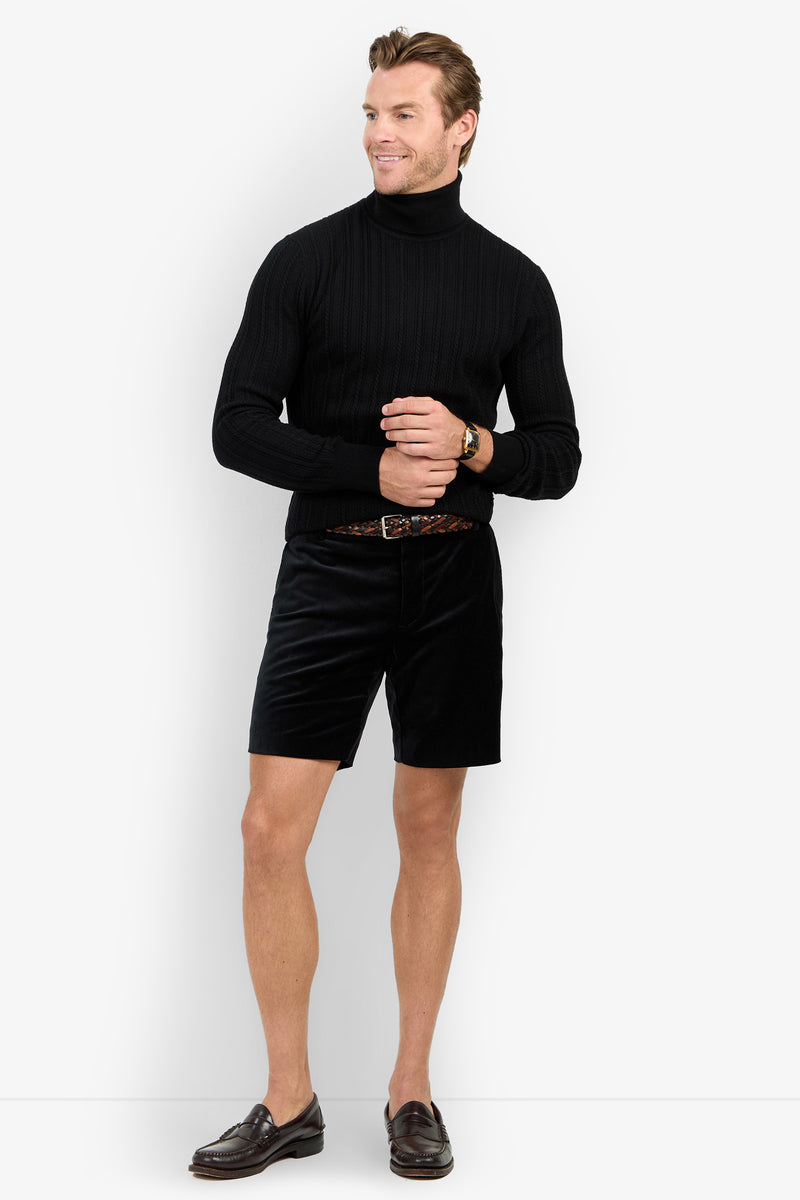 A man wears the Oberon Turtleneck Sweater - Merino Wool in black with black shorts, a brown belt, and brown loafers, smiling against a plain white background.
