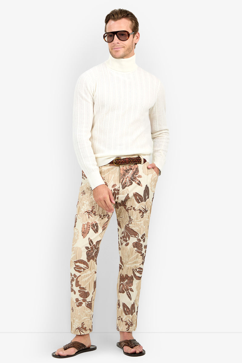 A man in an Oberon Turtleneck Sweater - Merino Wool, sunglasses, beige floral print pants, a brown belt, and flip-flops stands with one hand in his pocket against a plain background.