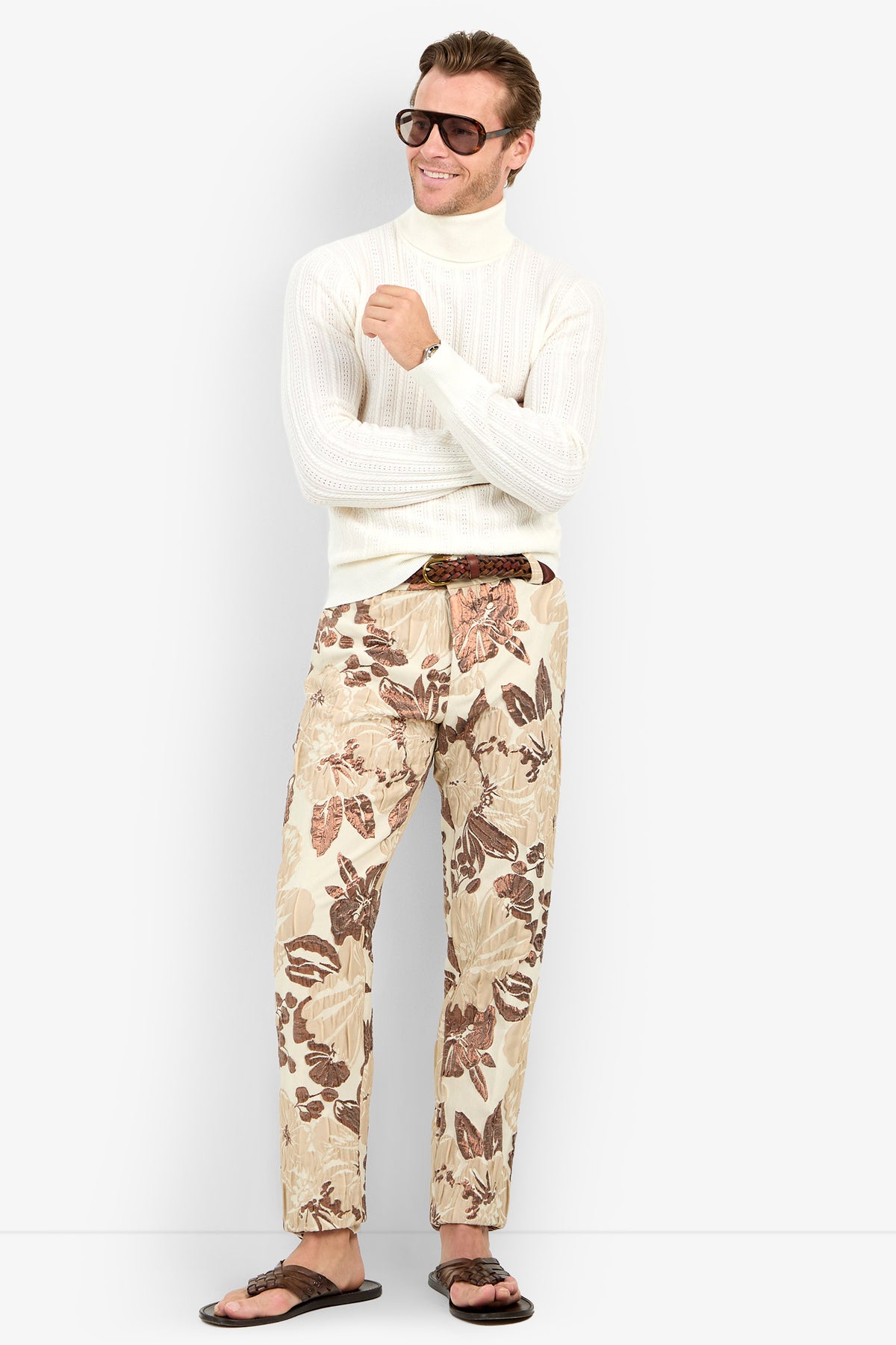 A man wearing sunglasses, the Oberon Turtleneck Sweater - Merino Wool, beige floral print pants, and brown sandals stands smiling with arms loosely crossed.