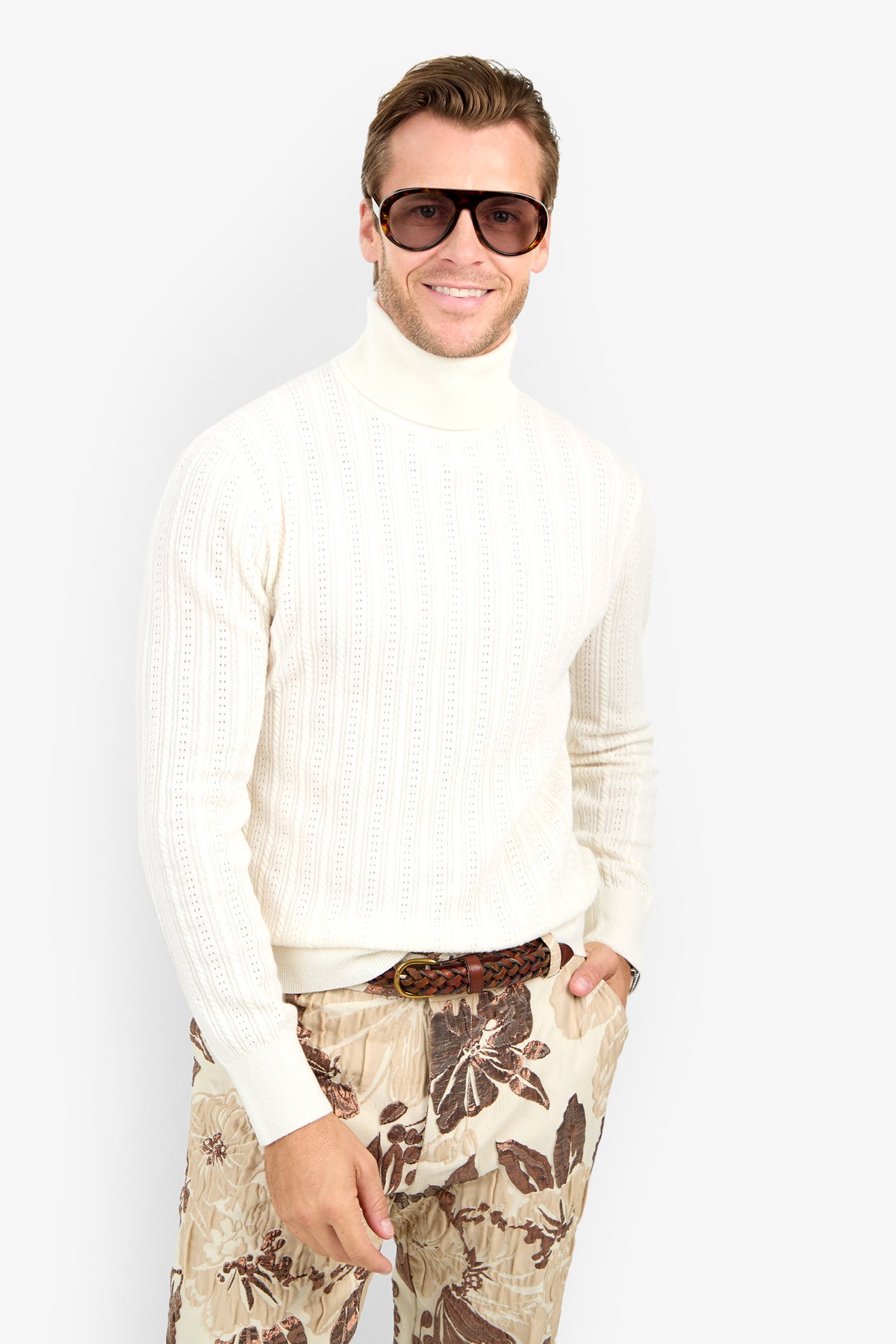 Man wearing Oberon Turtleneck Sweater - Merino Wool, large sunglasses, and beige floral pants, stands smiling against a plain white background.