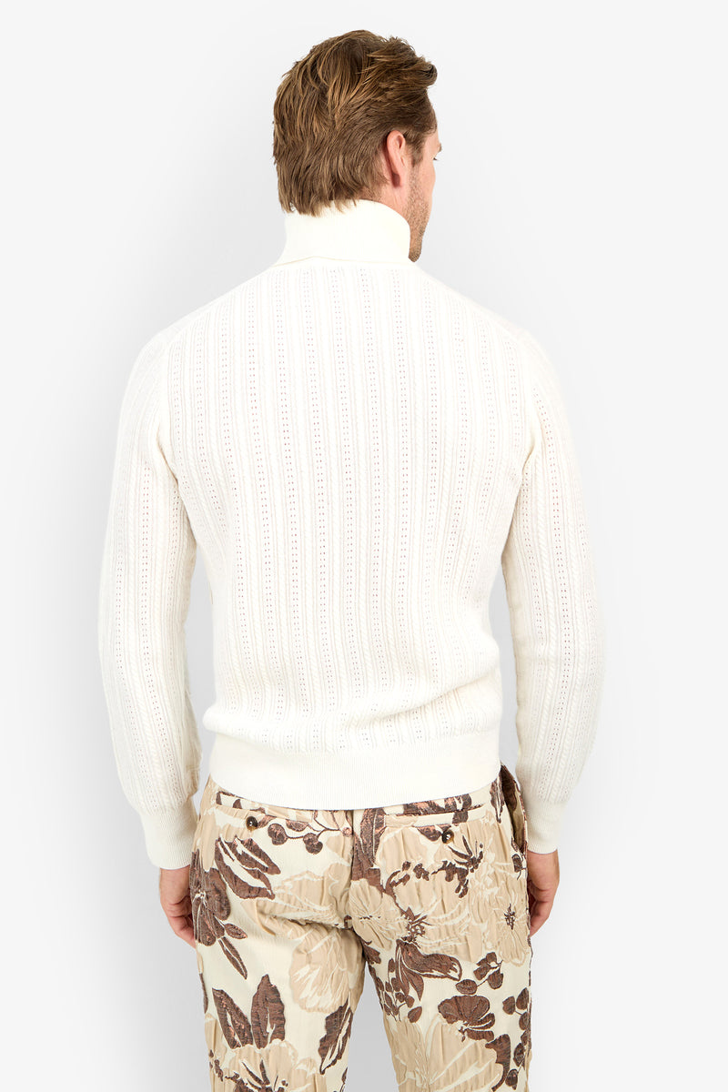 A man faces away, wearing the Oberon Turtleneck Sweater - Merino Wool in white with beige floral-patterned pants, set against a plain white background.