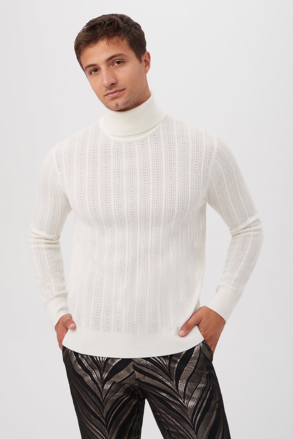A man wears the Oberon Turtleneck Sweater in Winter White merino wool and black metallic leaf pattern pants, standing against a plain light background with his hands in his pockets.