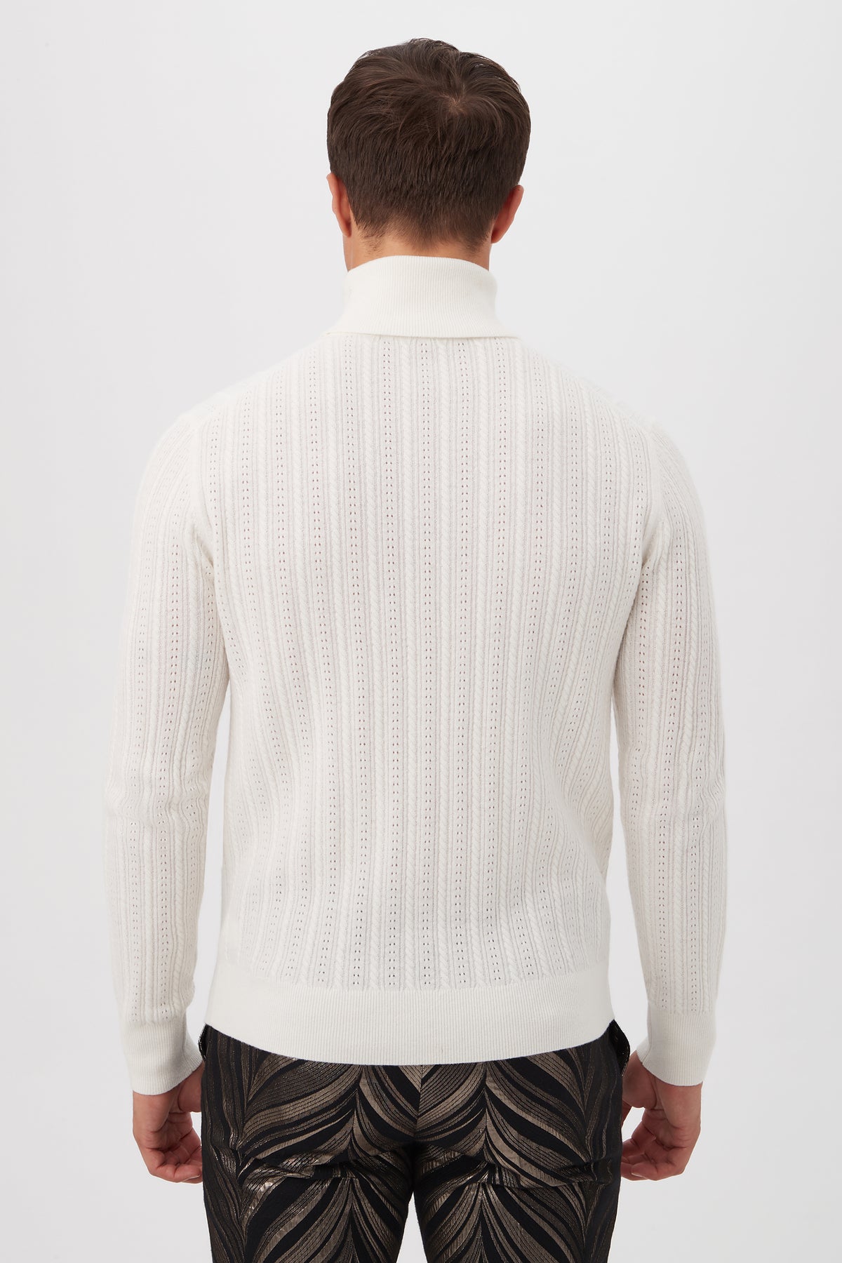 A man stands with his back to the camera, wearing the Oberon Turtleneck Sweaterâ€”crafted from merino woolâ€”in white textured knit, paired with dark abstract-patterned pants against a plain white background.