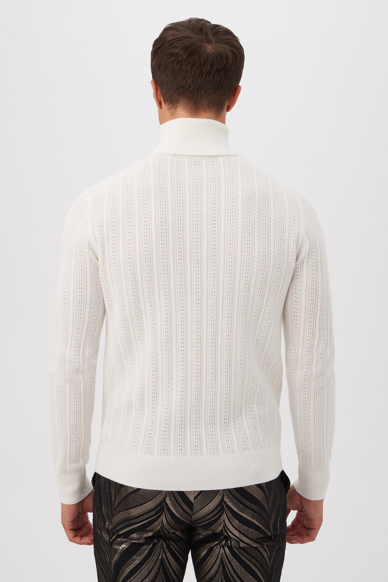 A man stands with his back to the camera, wearing the Oberon Turtleneck Sweaterâ€”crafted from merino woolâ€”in white textured knit, paired with dark abstract-patterned pants against a plain white background.