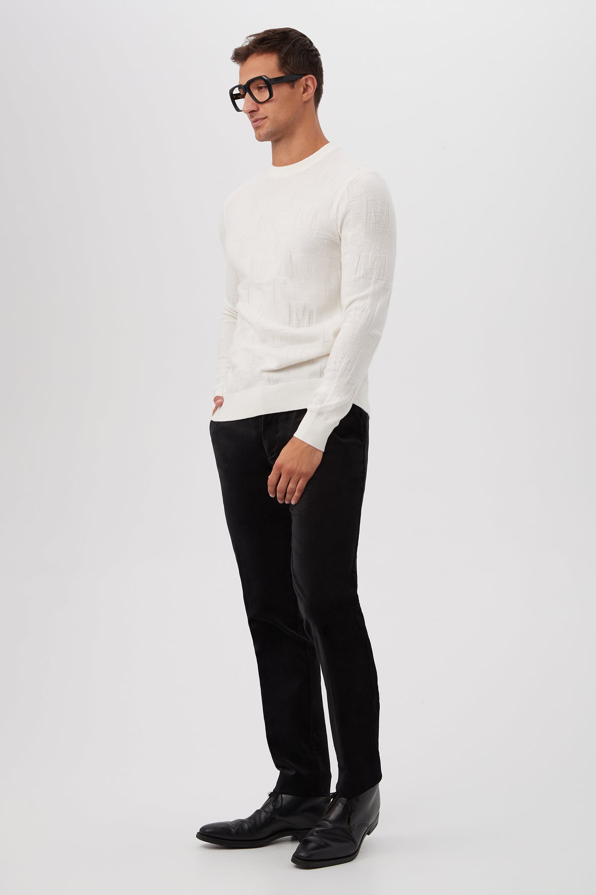 A man wearing black glasses, black pants, black shoes, and the Mr Turk Crewneck Sweater - Merino Wool stands against a plain white background with one hand in his pocket.