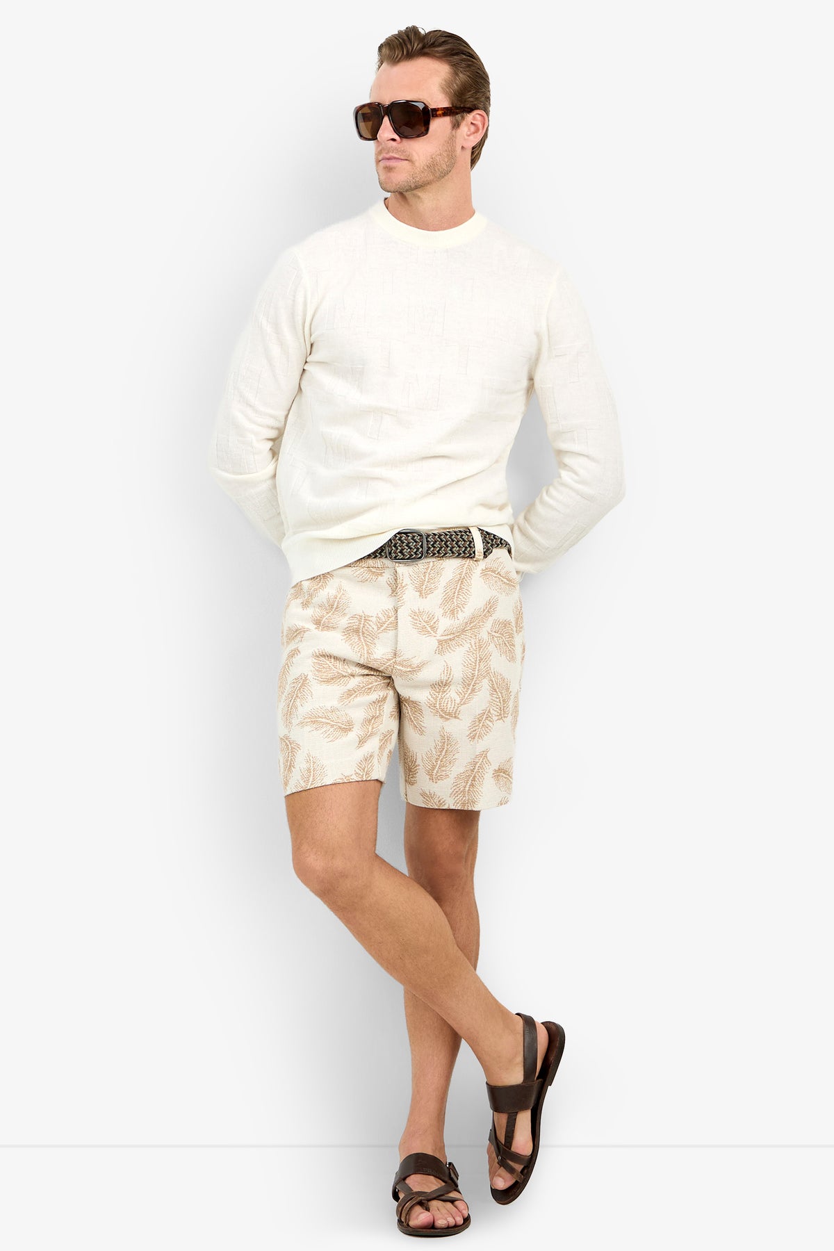 Man wearing sunglasses, the Mr Turk Crewneck Sweater - Merino Wool in white, patterned beige shorts, a black and white belt, and brown sandals stands with legs crossed against a plain white background.