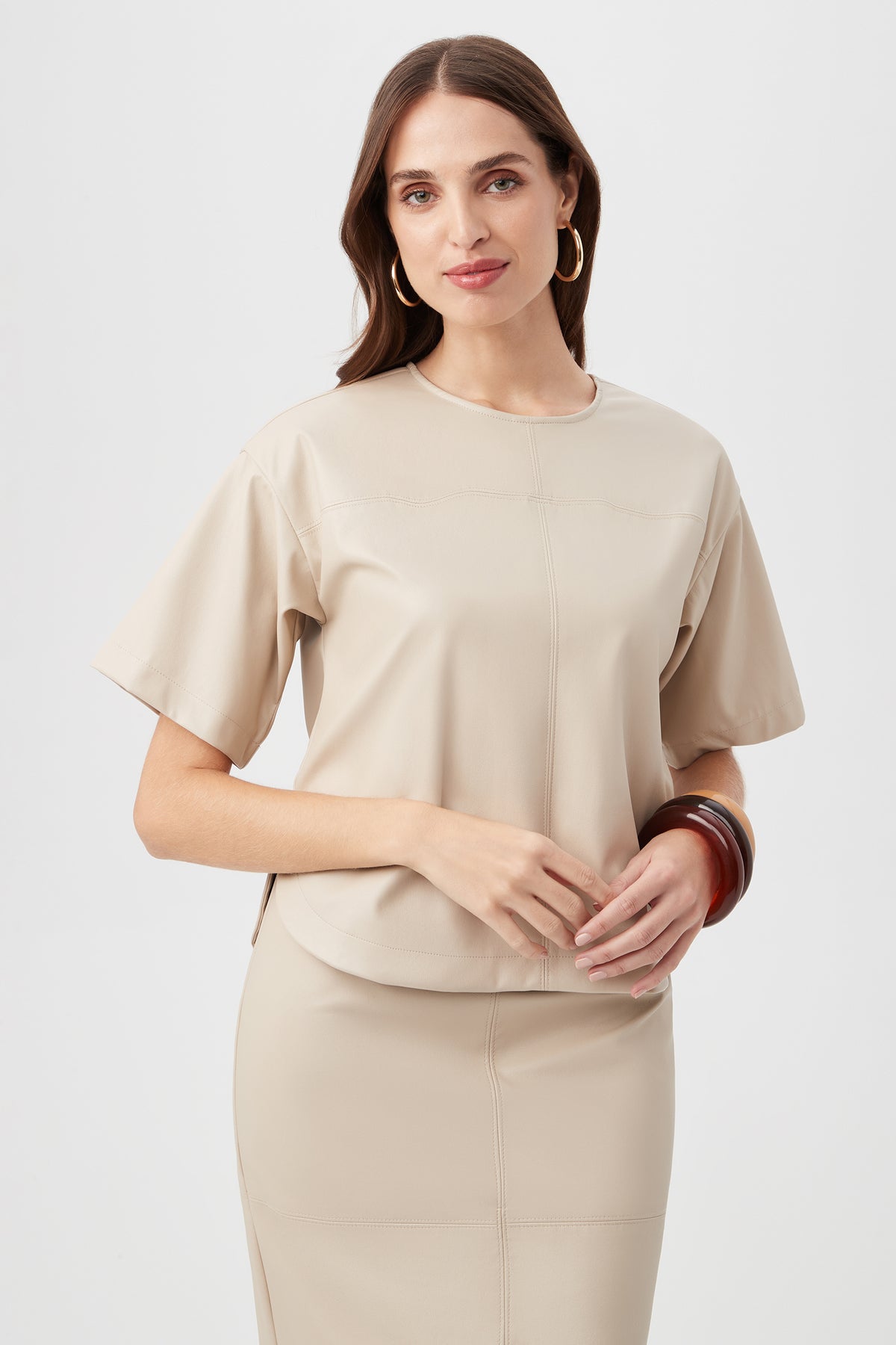 A woman with long brown hair stands against a light background, smiling softly. She wears the Moira Top in Chai with matching skirt, hoop earrings, and chunky bracelets on her left wrist.