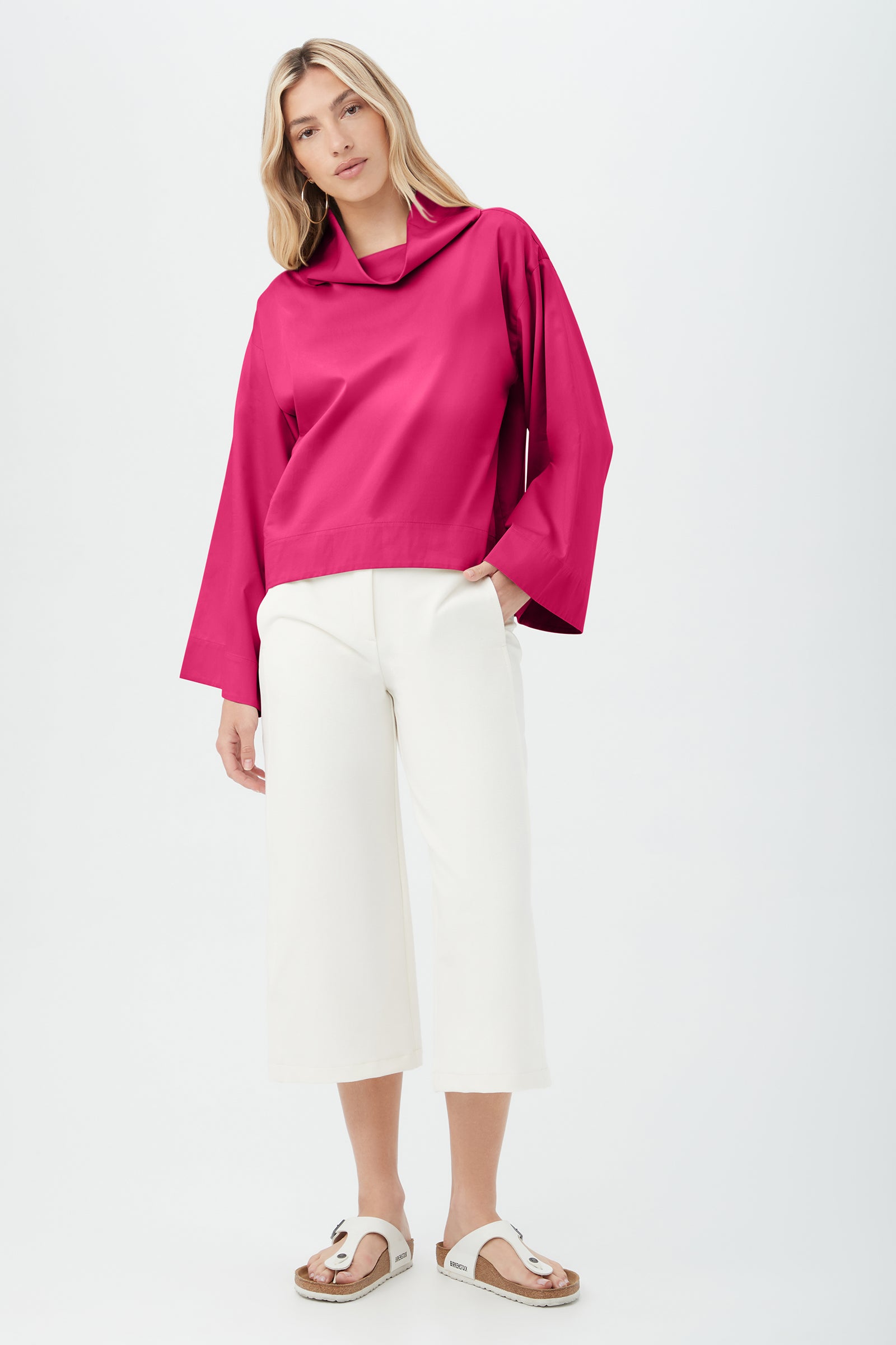 A woman stands against a plain background wearing the bright pink Tomie Top with a loose collar, wide white cropped pants, and white sandals. Her wavy hair is down, and she has one hand in her pocket.