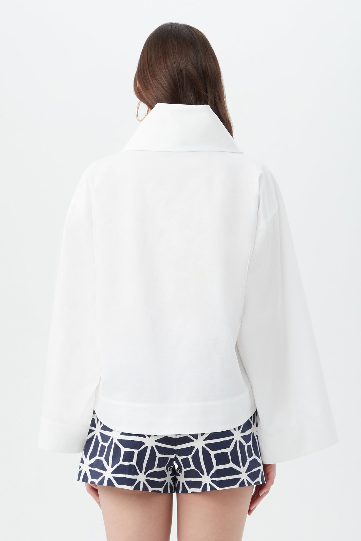 A woman with long brown hair faces away on a white background, wearing the Tomie Top—a white jacket with a wide collar—and navy blue shorts with a white geometric pattern.