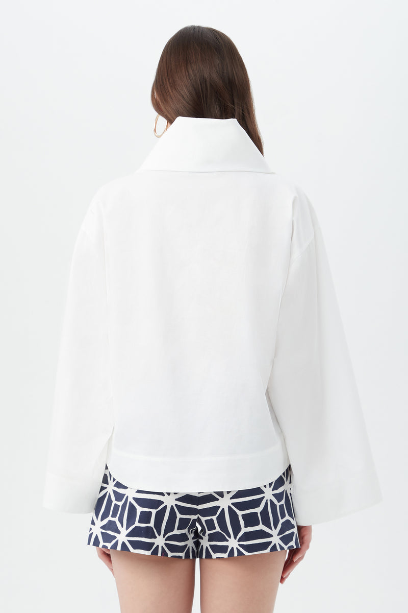A woman with long brown hair faces away on a white background, wearing the Tomie Top—a white jacket with a wide collar—and navy blue shorts with a white geometric pattern.