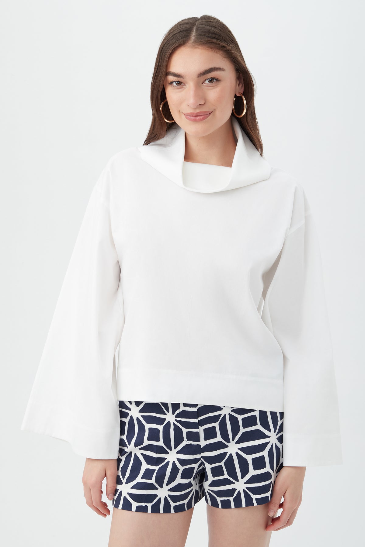A woman stands against a plain white background wearing the Tomie Top in white, a loose long-sleeve blouse with a wide collar, paired with navy blue shorts featuring a bold white geometric pattern.