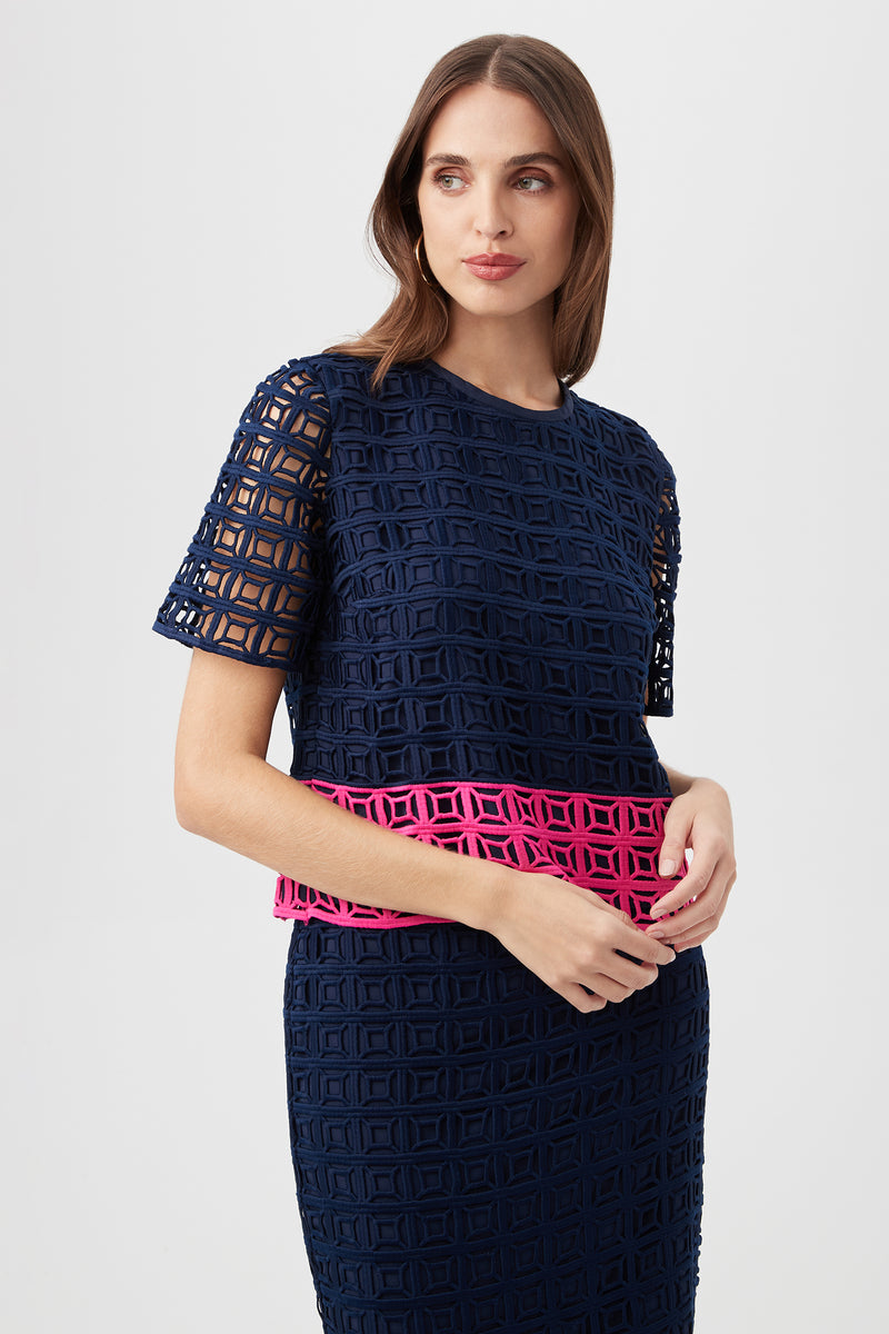 Woman wearing the Serilda 2 Top – Block Party Lace in Indigo/Petal Pink, featuring short sleeves, a geometric cutout pattern, and a bright pink waist band, posed against a plain light background.
