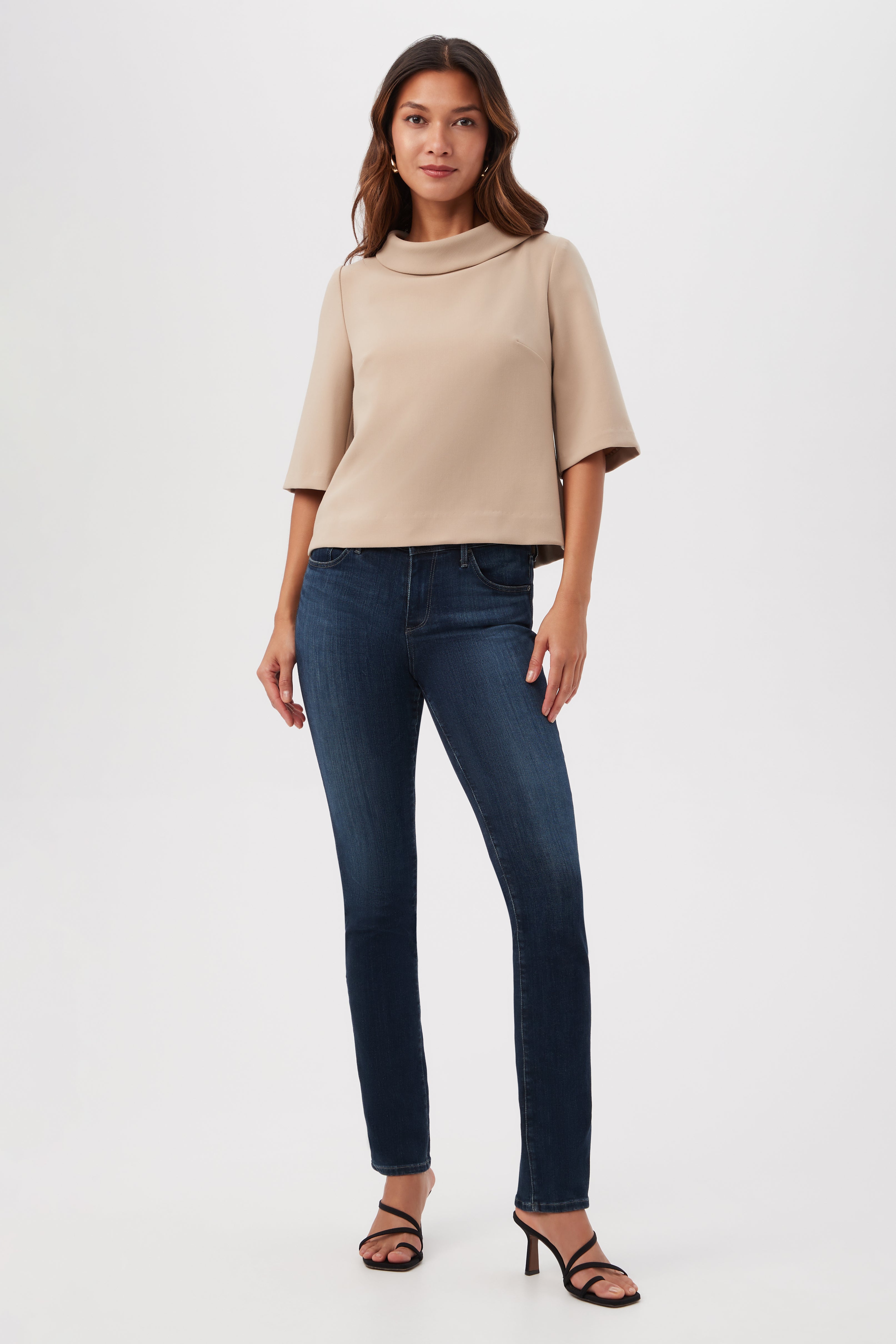 A woman with long brown hair stands against a white background wearing the Kailee Short-Sleeve Folded Collar Top in beige, dark blue fitted jeans, and black heeled sandals. She looks forward with a neutral expression.