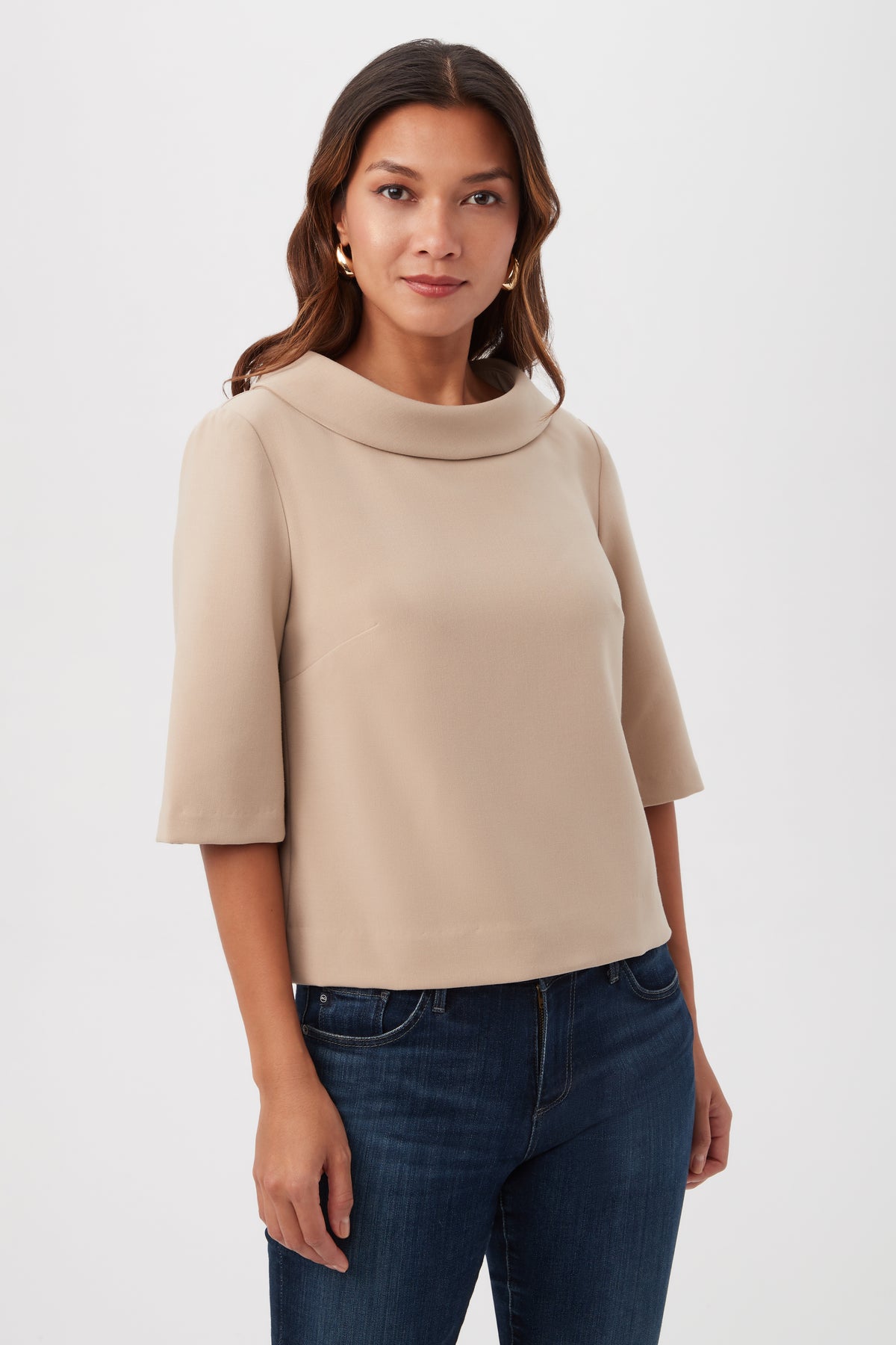 A woman with long brown hair wears the Kailee Short-Sleeve Folded Collar Top in Beach and blue jeans, standing confidently against a plain white background.
