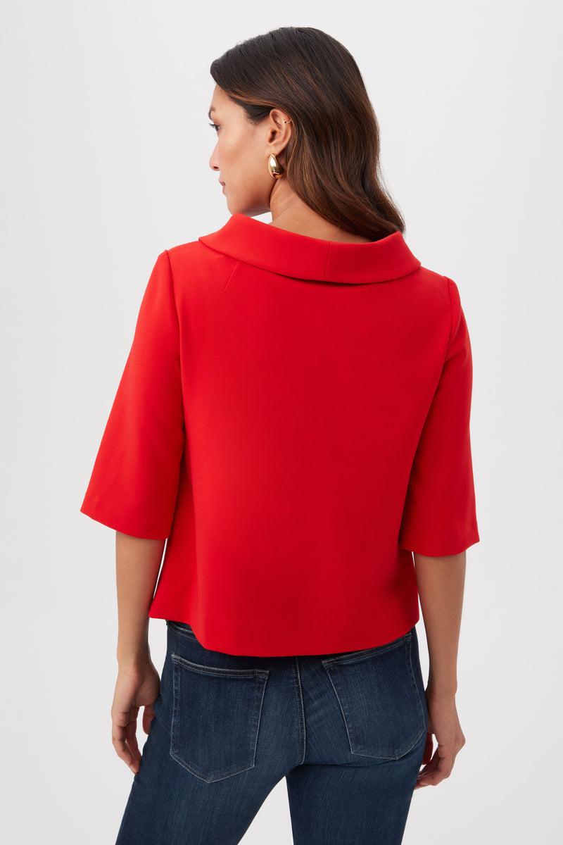 A woman with long brown hair, gold hoop earrings, and dark blue jeans wears the Kailee Short-Sleeve Folded Collar Top in bright red and stands facing away from the camera against a plain light background.