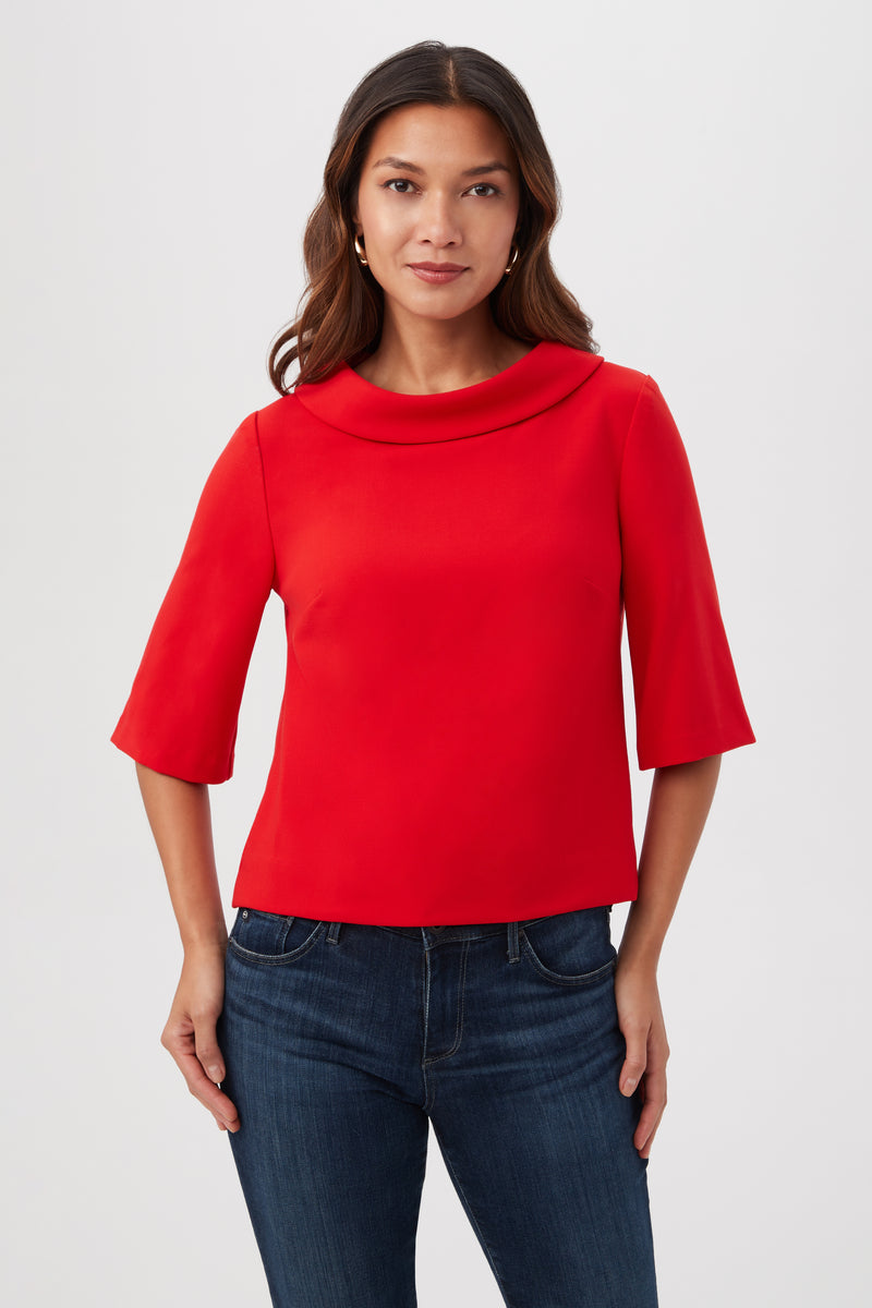 A woman with long brown hair wears the Kailee Short-Sleeve Folded Collar Top in Reina Red, paired with blue jeans, standing against a plain light background.