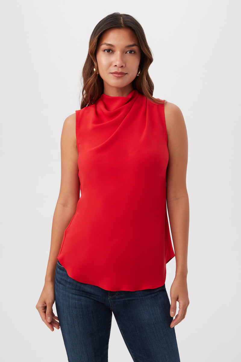 A woman with long brown hair models the Lenaya Sleeveless Cowl Neck Top in Reina Red, paired with blue jeans, standing against a plain white background.