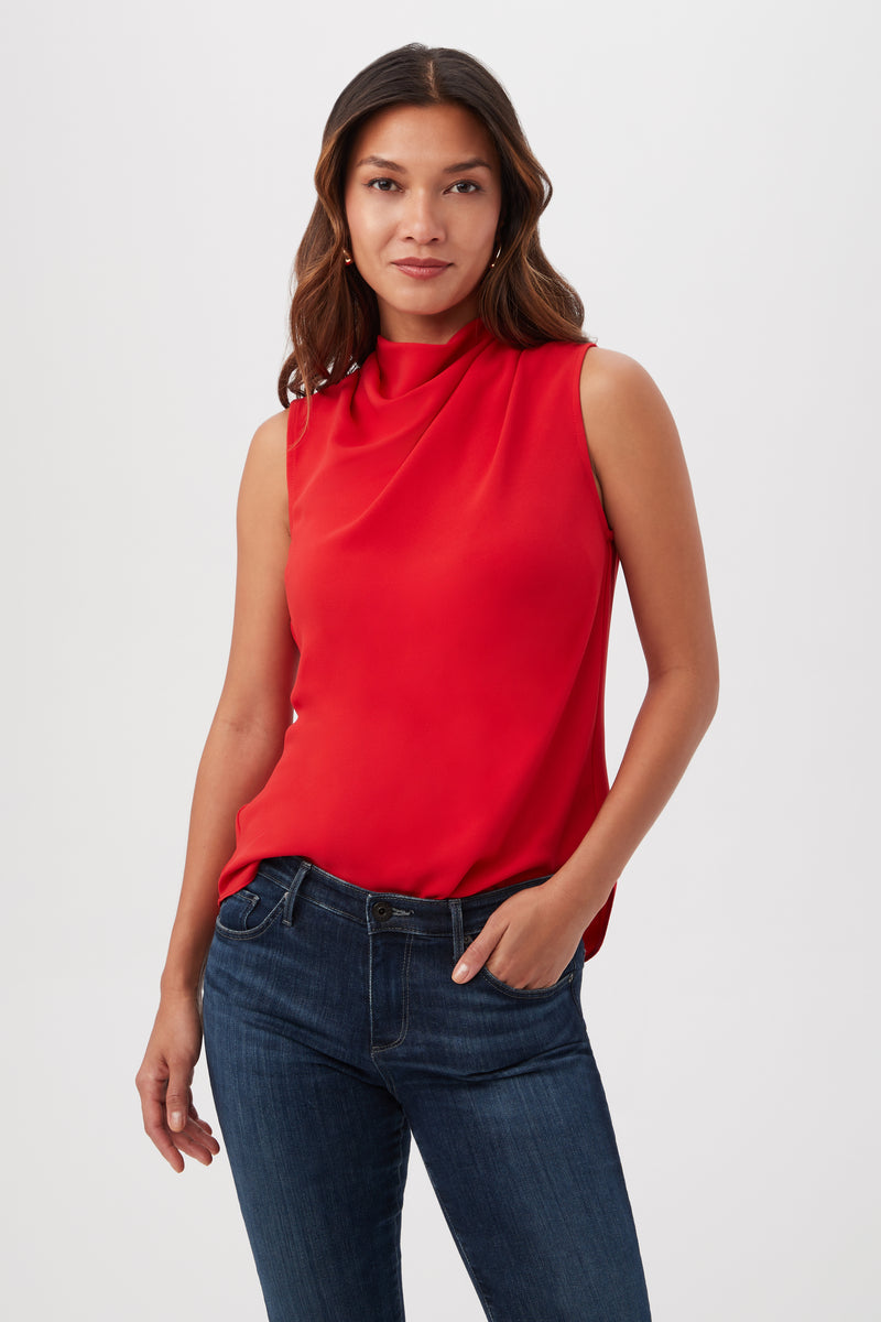Woman with long brown hair models the Lenaya Sleeveless Cowl Neck Top in red, paired with blue jeans. She stands against a plain white background, one hand in her pocket and a slight smile on her face.
