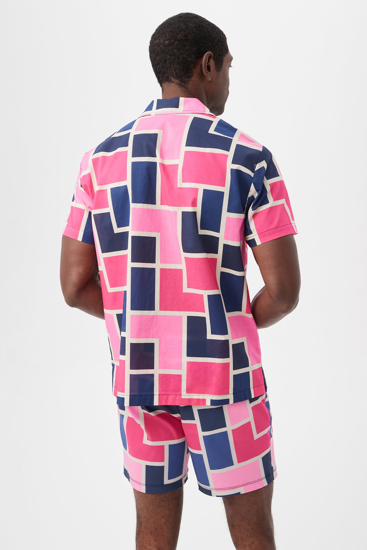 A man is seen from behind wearing the Felix Camp Shirt - Block Party, a bright short-sleeve set featuring a geometric pattern of pink, navy blue, and white rectangles on a white background.