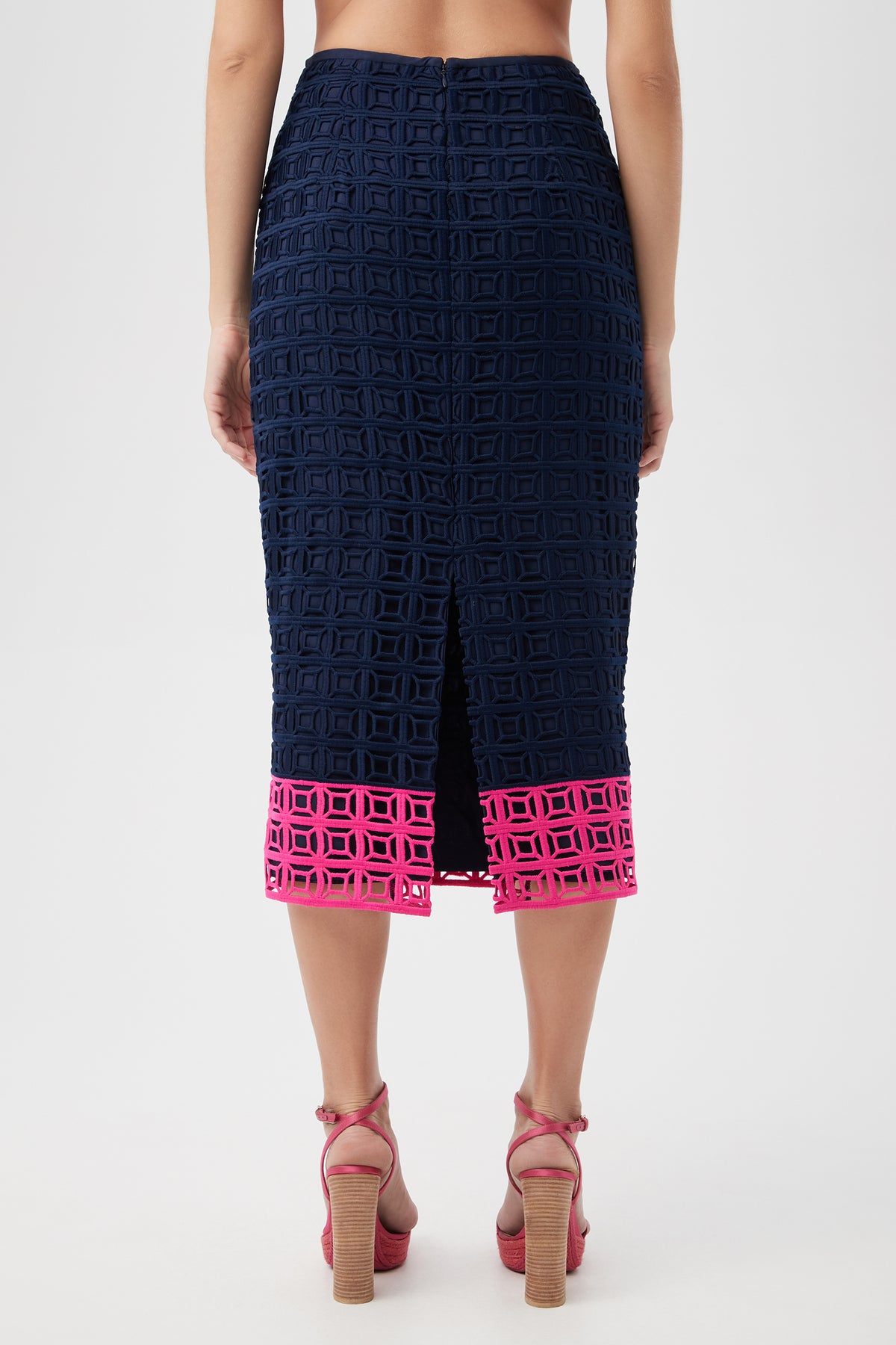 A woman is shown from the back wearing the Dita 2 Midi Pencil Skirt - Block Party Lace, featuring navy geometric cutout lace, a center slit, and a vivid pink cutout border at the hem. She pairs it with pink high-heeled sandals.