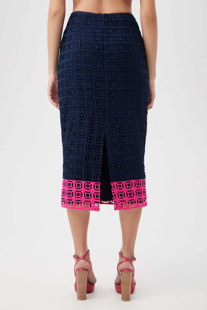 A woman is shown from the back wearing the Dita 2 Midi Pencil Skirt - Block Party Lace, featuring navy geometric cutout lace, a center slit, and a vivid pink cutout border at the hem. She pairs it with pink high-heeled sandals.