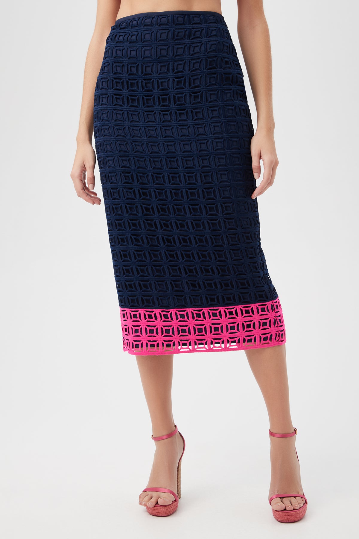 A person wearing the Dita 2 Midi Pencil Skirt - Block Party Lace in Indigo/Petal Pink, featuring a geometric cut-out design and petal pink border, paired with matching pink high-heeled sandals.